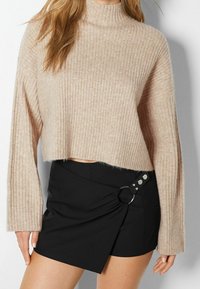 Beige-värinen ribbineulepaita, jossa on korkea kaulus ja pitkät hihat, yhdistettynä mustaan kietaisuhameeseen, jossa on hopeanvärinen rengasdetaili.