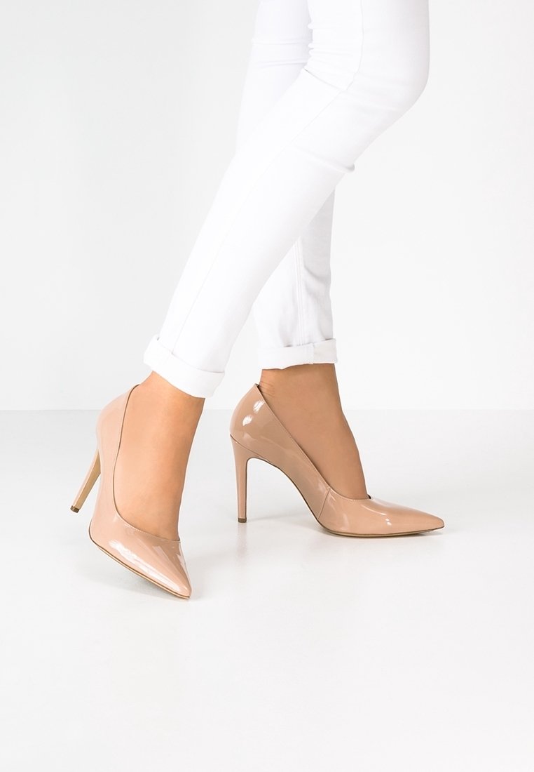 Jambes portant un pantalon blanc retroussé et des escarpins à bout pointu en cuir verni nude sur un sol blanc.