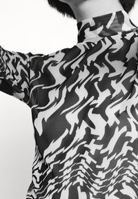 Primo piano di una persona che indossa un top a collo alto con motivi astratti ondulati in bianco e nero e capelli corti scuri.