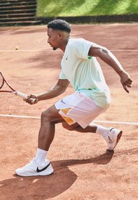 Tennisspieler in einem mintgrünen Kurzarmhemd und weißen Shorts mit gelben Akzenten, der auf einem roten Sandplatz mit einem Schläger spielt und weiße Sneaker trägt.