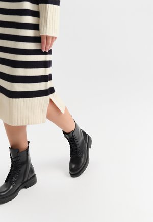 Czarne skórzane buty wojskowe na sznurówki z grubą podeszwą, w zestawieniu z kremowo-granatową sukienką z prążkowanej dzianiny z bocznymi rozcięciami.