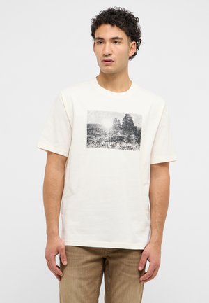 Junger Mann mit lockigem Haar, der ein weißes T-Shirt mit einem schwarz-weißen Felslandschaftsprint und braune Hose vor einem schlichten Hintergrund trägt.