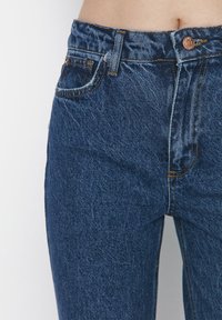 Jeans in denim blu scuro con vita alta, caratterizzati da una leggera scoloritura, bottoni color rame e design standard a cinque tasche.