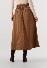 Vrouw in een lange bruine rok met een elastische tailleband, gecombineerd met donkerbruine haklaarzen en een beige geribbelde lange mouwtop.
