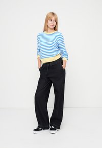 Modro-bel pulover s črtami z rumenimi poudarki, združen z črnimi širokimi hlačami in črnimi supergami. Priložnostna, udobna obleka.