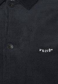 Sort fløjlsskjorte med en tekstureret overflade, udstyret med knaplukning og et broderet "VOLCOM" logo på venstre brystlomme.