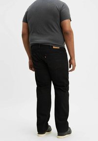 Jeans in denim nero con taglio a gamba dritta, caratterizzati da tasche posteriori e un piccolo tag in pelle con il marchio vicino alla vita. Indossati con una maglietta grigia.