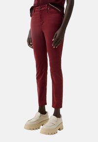 Burgundy slim-fit pants with a smooth texture, featuring a front zip and button closure. Paired with chunky white loafers with a fur trim.