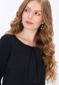 Sötétkék gyapjúkeverék felső, kerek nyakkivágással, hosszú ujjak, elöl masnival díszítve, és dekoratív gombbal. Sima textúra.