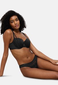 Γυναίκα με σγουρά μαλλιά ξαπλωμένη, φορώντας ένα μαύρο σουτιέν με λεπτομέρειες δαντέλας και αντίστοιχα μαύρα εσώρουχα με δαντελένιες λεπτομέρειες σε απλή φόντο.