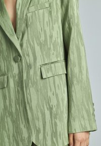 Blazer oversize verde chiaro con motivo testurizzato simile alla venatura del legno, con chiusura a bottone singolo e tasche frontali.