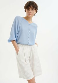 Helles blaues, locker sitzendes Blusenshirt mit einem runden Halsausschnitt und perlenartigen Knöpfen auf der Vorderseite; kombiniert mit weißen Shorts mit Nadelstreifen.