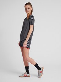 Jeune femme debout de profil, portant une tenue de sport sombre avec des motifs en chevrons et des baskets blanches avec des détails orange.