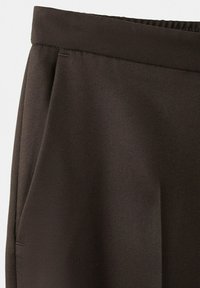 Temno rjavo teksturirano blago v bližnjem posnetku, ki prikazuje pas in stranski žep oblačila, verjetno hlač ali kratkih hlač.