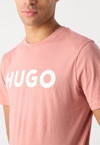 Šviesiai rožinė marškinėliai, pagaminti iš minkštos medvilnės, su ryškiu baltu "HUGO" logotipu ant krūtinės ir klasikinio apykaklės dizaino. Trumpomis rankovėmis.