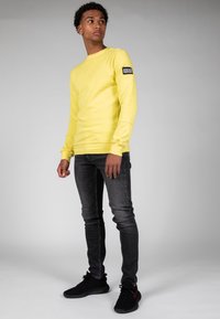 Sudadera amarilla de manga larga con puños acanalados y un parche de logo en la manga izquierda, combinada con jeans oscuros de corte ajustado y zapatillas negras.