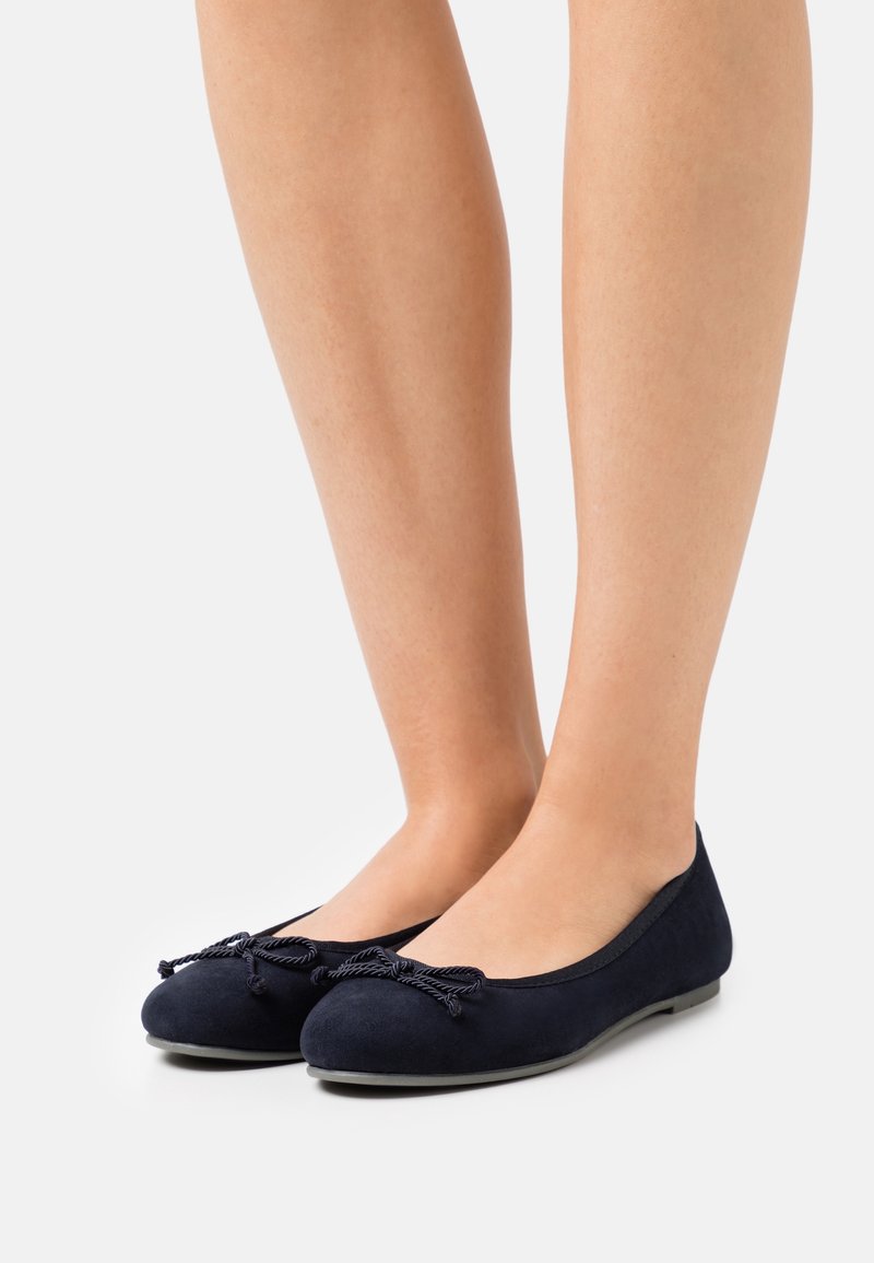 Jambes portant des ballerines bleu marine avec un nœud en corde sur des semelles grises sur fond blanc uni.