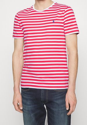 Homme portant un t-shirt à manches courtes à rayures horizontales blanches et rouges, et un jean bleu foncé, debout devant un fond uni.