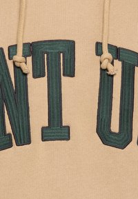 Beżowy dres z kapturem z zielonym haftowanym napisem "VENTU". Posiada kremowe sznurki i teksturowane wykończenie.