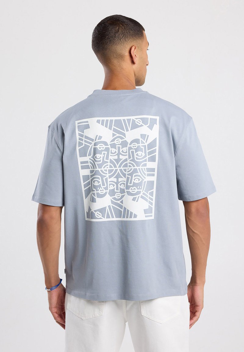 T-shirt en coton gris clair à manches courtes présentant un grand motif géométrique blanc au dos, représentant des visages et des formes abstraites.