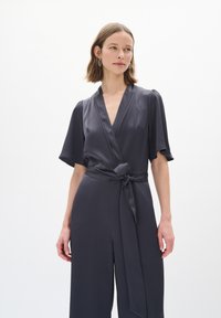 Bodysuit avvolgente grigio scuro con maniche corte, cintura legata e texture liscia. Presenta un colletto a V e una design con gambe larghe.
