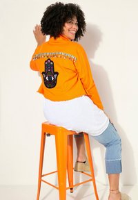 Orange avkortad jacka med färgglada dekorationer och ett hamsa-handdesign på baksidan. Modellen sitter på en orange metallpall.