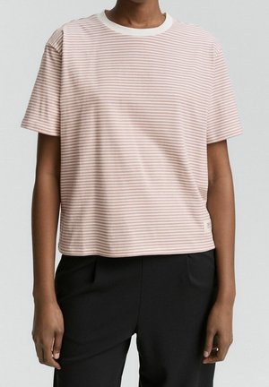 Personne portant une chemise à manches courtes rayée rose clair et blanche avec un col blanc et un pantalon noir, debout devant un fond uni.
