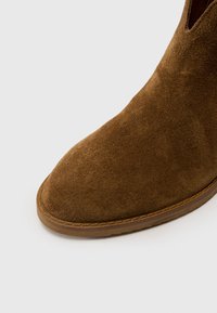 Bottine en daim marron à bout pointu et semelle en caoutchouc plate. La tige présente une texture lisse sans accents ni motifs saillants.