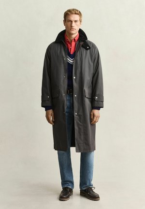 Homme portant un long manteau sombre par-dessus un pull bleu marine à rayures blanches, une chemise rouge, un jean bleu et des chaussures marron foncé, debout devant un fond uni.