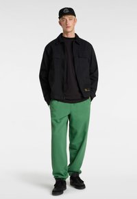 Zwarte canvas jack met twee borstzakken, zwart t-shirt en groene sweatpants. Zwarte pet met wit logo en zwarte sneakers.