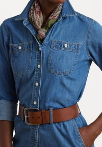 Chemise en denim bleue avec un col, deux poches poitrine et une fermeture à boutons ; associée à une ceinture en cuir marron et un foulard floral vert.