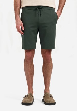 Homme portant un short vert foncé à cordon et des chaussures beige en daim sans lacets, debout sur un fond blanc.