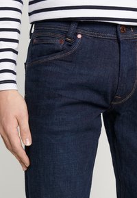 Mörkblå denimjeans med kontrasterande orange sömmar, femficksdesign och kopparfärgad hårdvara. U-benägen vid benslutet.