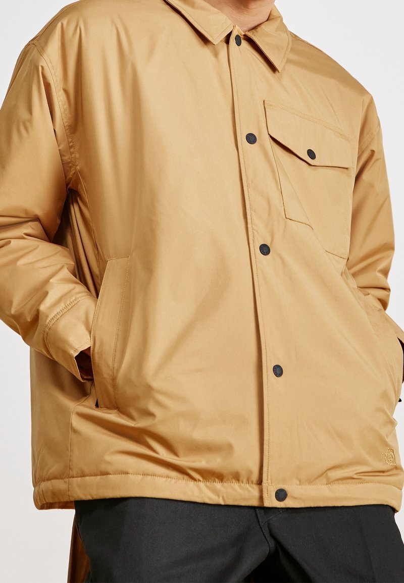 Veste beige à boutons-pression noirs, une poche poitrine avec rabat, poches latérales, portée par une personne les mains dans les poches.