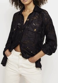 Camicia nera in pizzo con bottoni, dal motivo testurizzato, due tasche frontali e maniche arrotolate. Indossata su jeans bianchi a vita alta.