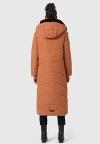 Long, orange puffer coat with a hood and black faux fur trim, featuring chevron quilting and a logo patch at the hem. Worn over black pants and boots.
