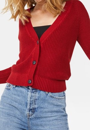 Kobieta ubrana w czerwony sweter zapinany na guziki, na czarnym topie i jasnoniebieskich, wysokich jeansach, pozująca z lekko uniesioną jedną ręką.
