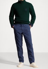 Vihreä neulekauluspaita, jossa on ribbikuvio, yhdistettynä tummansinisiin chinopantseihin ja vaaleanpunaisiin lenkkitossuihin. Kello näkyy vasemmassa ranteessa.