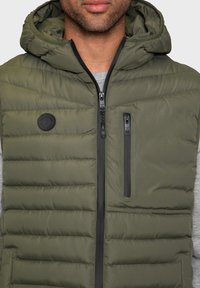 Doudoune sans manches vert olive, dotée d'une fermeture éclair frontale, d'une capuche et de motifs matelassés horizontaux. Comprend un petit patch logo et une poche latérale zippée.