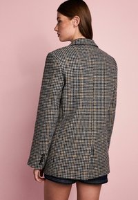 Houndstooth raštuotas švarkas tamsių ir šviesių atspalvių, su prigludusia siluetu, iškirptėmis atlapais ir sagomis ant rankovių. Pavaizduota nugarinė pusė.
