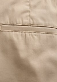 Beżowa tkanina z poziomym szwem kieszeni na listwę i widocznym przeszyciem, ukazująca fakturę i lekkie zmarszczki na materiale.