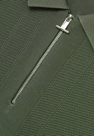 Îmbrăcăminte cu fermoar de culoare verde khaki, cu un design texturat, în dungi, și un fermoar metalic. Prezintă un guler în picioare și accente din țesătură netedă.