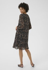 Blaues, transparentes Kleid mit floralem Muster, drei Viertelärmeln und einem rüschierten Saum. Modell trägt schwarze flache Schuhe und steht vor einem neutralen Hintergrund.