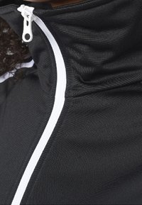 Svart zip-up jacka med hög krage, som har en vit dragkedja och texturerat tyg. Designen inkluderar kontrasterande kantband längs framsidan.