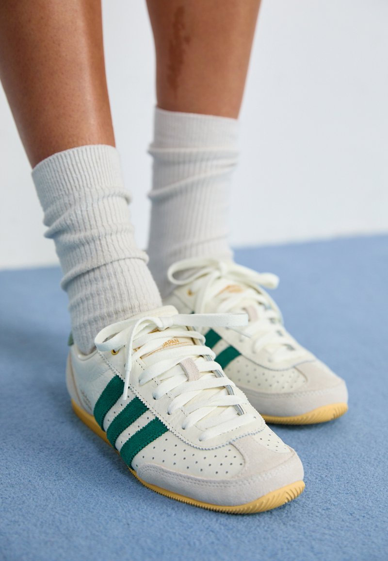 Ténis brancos com riscas verdes, detalhes em camurça perfurada, atacadores planos e uma sola de borracha texturizada, combinados com meias cremosas de malha.