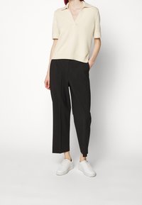 Krémové polo tričko s krátkým rukávem a V-výstřihem, které je kombinováno s černými širokými kalhotami a bílými teniskami. Materiál vypadá hladce a lehce.