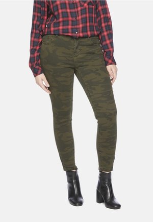 Persoon draagt groene camouflage skinny broek, rood en marineblauw geruit overhemd en zwarte enkellaarsjes met hak, staand tegen een witte achtergrond.