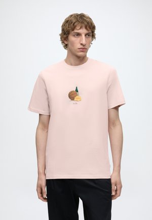 Jeune homme portant un t-shirt rose clair avec un motif de basketball, bouteille de champagne et croissant, associé à un pantalon noir, fond neutre.