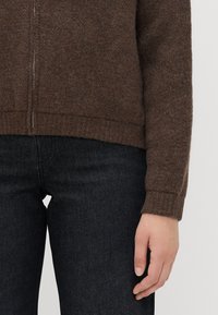 Brun ulltröja med dragkedja, ribbad nederkant och långa ärmar, i kombination med mörka jeans. Texturerad tyg synligt.