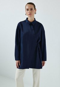 Cămașă bleumarin cu nasturi, cu detaliu de cravată, mâneci lungi și guler, confecționată dintr-un material neted. Purtați-o cu pantaloni de culoare deschisă.